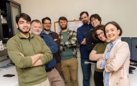 A group of eight people, two women and six men, stand together in a lab space with shelves on one side and a bank of computers on the other. All are smiling, facing the camera, with their arms folded across their chests.
