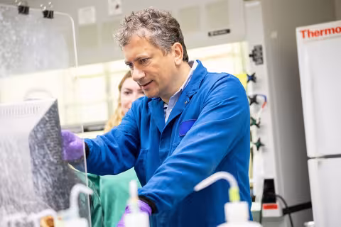 A man wearing a blue lab coat and purple gloves points to a computer screen in a brightly lit lab. 