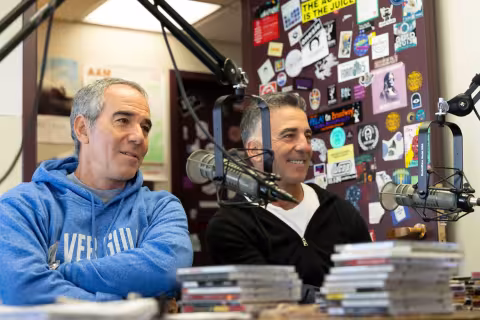 Two men sit by a microphone inside of a Radio studio, on the left a man wears a bright blue sweatshirt and on the right a dark sweatshirt.