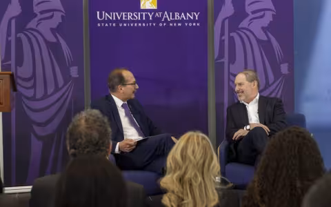 Two men in suits chat unfront of an audience, with a puple "University at Albany" banner behind them.