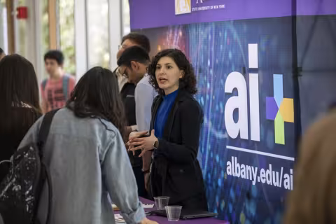 A person standing and speaking with with someone in front of a banner with UAlbany's AI+ logo behind them.