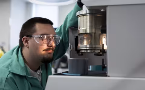 A man wearing glasses, protective goggles and a green lab coat handles a lab instrument that features a metal cylinder containing a lit glass chamber.  