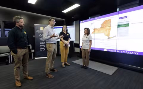 Governor Kathy Hochul stands with Nick Bassill and other New York emergency management leaders in front of a weather map inside the xCITE lab.