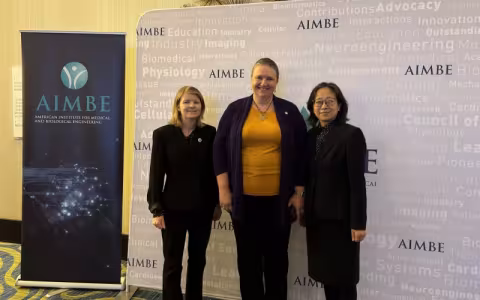 Three women smile for a photo, standing against a backdrop printed with the name “AIMBE” in purple, with a collection of science-related words in very faint print. To their left is a pull-up poster that says “AIMBE American Institute for Medical and Biological Engineering”. The woman on the left is wearing a black blazer and slacks. The woman in the center is wearing an orange top with black cardigan. The woman on the right is wearing black square rimmed glasses and a polka dot blouse under a black blazer. 