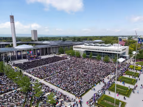 An aerial shot of the University at Albany main campus with a large crowd of people in the central plaza with a blue sky and large carillon in the background.