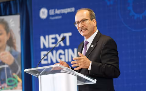 A man in a black suit, purple tie and glasses speaks at a podium on a stage in front of a screen that reads "Next Engineers"