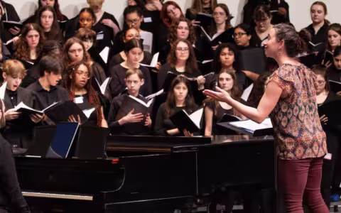 Young people dressed in all black are holding music books and singing on a stage with a piano and conductor.