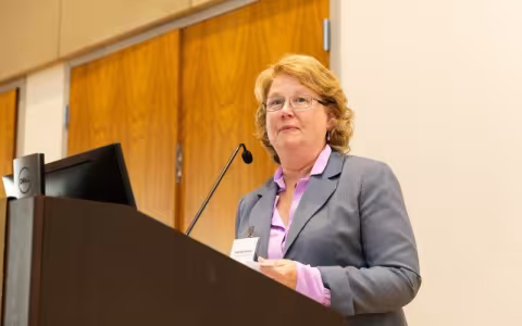 A woman in a gray jacket and purple shirt stands at a microphone on a lecturn