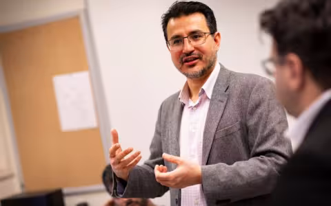 A man with dark hair and a gray blazer and glasses speaks animatedly in a brightly lit classroom. 