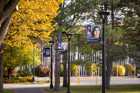 Five banners hang from light posts on a campus path on a sunny fall day. The first banner includes the University at Albany logo and the Community Engaged Scholars logo. The next three the banners include headshots of community engaged faculty memberr. The fifth banner includes a QR code.