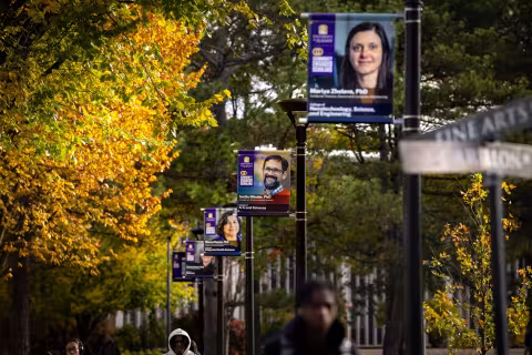Four banners hang from light posts on a campus path on a sunny fall day. Three of the banners include headshots of community engaged faculty members and the fourth banner includes a QR code.
