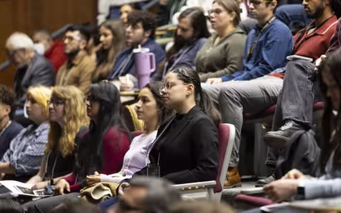 Rows of people sit in auditorium-style seats, listening intently to a speaker at the front of the room.
