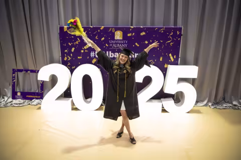 A UAlbany graduate student stands in cap and gown with arms upraised in front of a lit sign that reads 2025