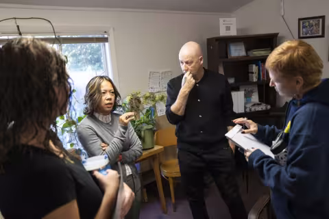 Four people stand mid-discussion in a white-painted room with a bookshelf, plants and a window behind them.