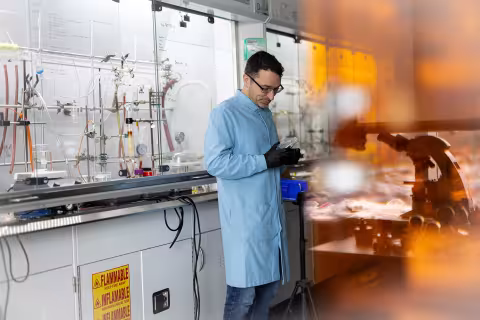 A man wearing a pale blue lab coat and black gloves examines a beaker in a chemistrylab.