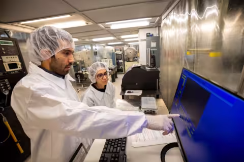 A man in a white cleanroom "bunny suit" gestures to a blue computer screen while a woman in a similar suit looks on.