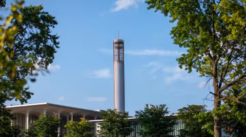 The UAlbany carillon, framed by lush green trees and photographed against a bright blue sky.