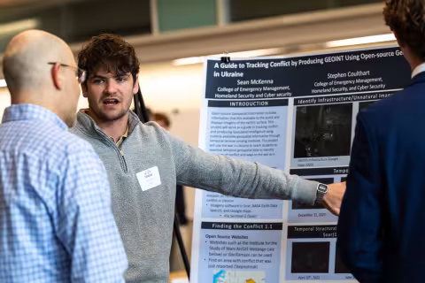 A student presenter gestures toward his research poster while speaking with a Showcase attendee about tracking conflict in Ukraine.