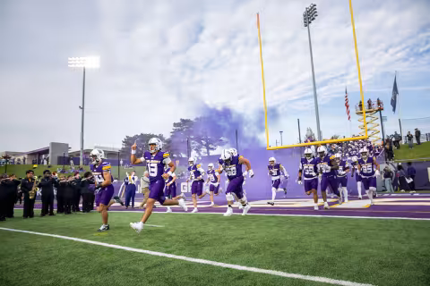 UAlbany football players running through purple smoke on to the field for the 2025 Homecoming game.
