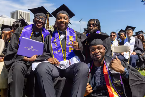Three students in full regalia smiling and posing with their diplomas at commencement.