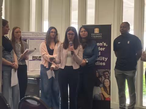 Five students standing behind a student talking on a microphone.