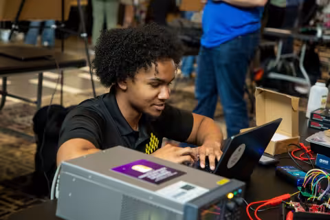An engineering student crouches to work on a laptop placed on a table where wires connect to a CPU.