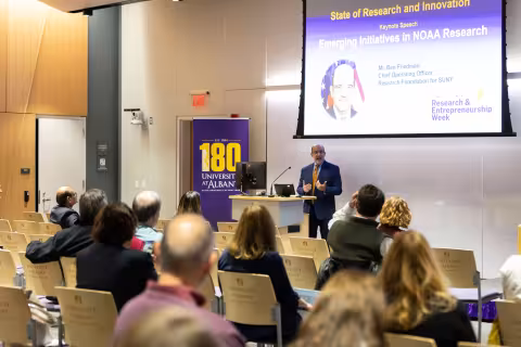 Benjamin Friedman, Chief Operating Officer of the Research Foundation for SUNY, speaks in front of a seated crowd at R&E Week 2024. The slide projected at a screen at the front of the room includes the title, "State of Research and Innovation, Keynote Speech, Emerging Initiatives in NOAA Research."