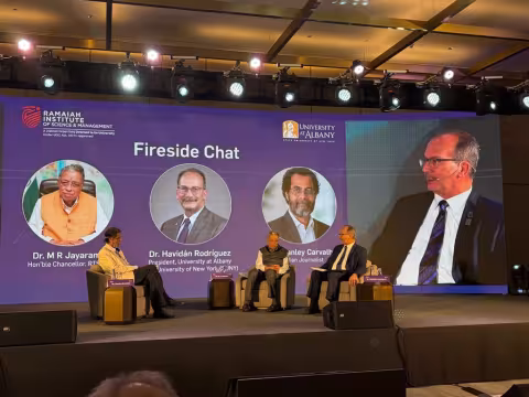 Three officials from UAlbany and RISM sit on stage and engage in a panel discussion. Behind them, a screen shows their names, titles and photos, as well as the words, "Fireside Chat."