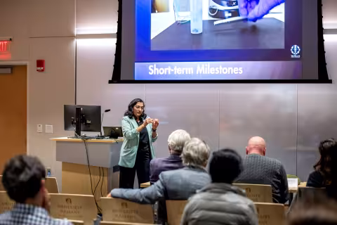 A presenter gestures while speaking to a seated audience at R&E Week 2025. The slide displayed via projector includes the title "Short-term Milestones."