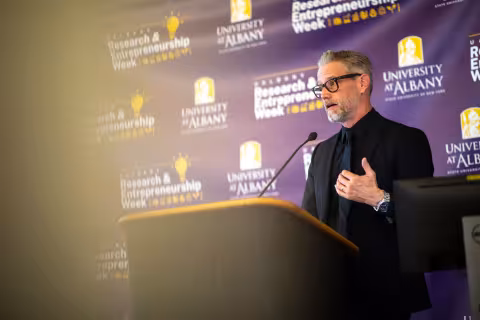 A researcher stands at a podium and speaks during R&E Week 2023. He stands in front of a purple backdrop with the event logo.