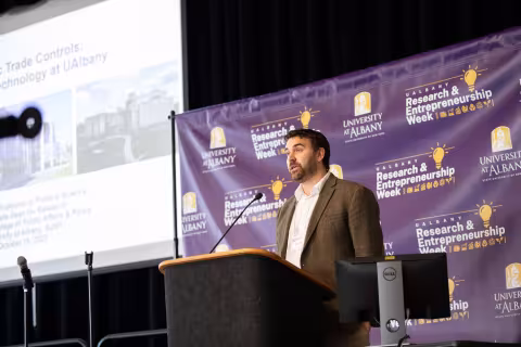 A researcher speaks from a podium in front of an R&E Week 2023 backdrop. The slideshow being projected on the screen mentions trade controls.
