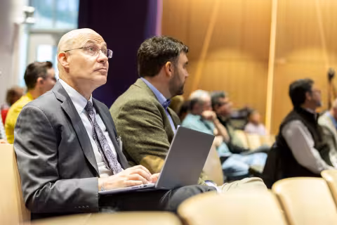 An R&E Week 2024 attendee sits in the audience, listening intently and taking notes on a laptop.