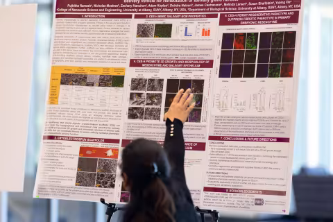 A researcher sits at a table and gestures behind her towards a research poster at R&E Week 2024.