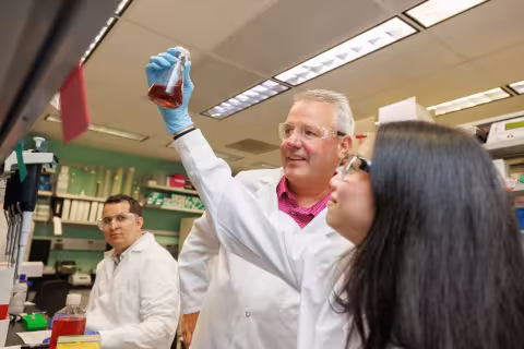 A young woman wearing lab goggles and a white lab coat holds up a beaker containing a red liquid. A man wearing a white lab coat and plastic protective goggles stands behind her, observing the liquid inside the beaker. A man wearing a white lab coat and goggles works at the lab bench behind them. 