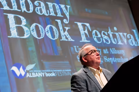 A man speaks at a podium with the words Albany Book Festival behind him on a large screen.