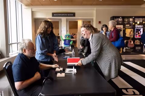 A man is seated signing books as several people line form to get autographs and a woman leans over a book she has placed in front of him.