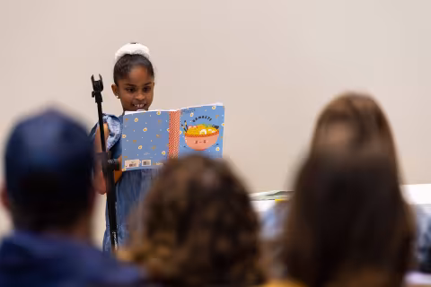 A young girl with a white bow in her hair reads from a book at a microphone in front of an audience.