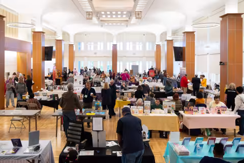 A large group of people move between aisles of people seated with their books during a book show in an auditorium