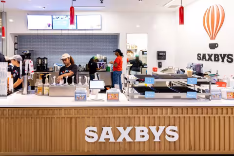 Three students work behind the counter of a coffee eatery with the name SAXBYS in capital letters underneath the counter