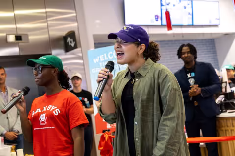 Two young women, one in a red shirt and another in a gray buttoned shirt and hat, sing in front of a new cafe opening inside the Massry School of Business