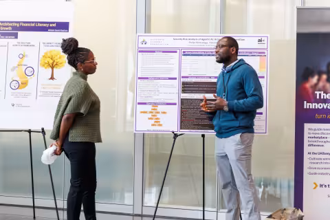 A presenter stands in front of a research poster and gestures while speaking with an attendee during the 2026 AI Plus Symposium.