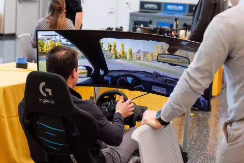 A person sits in a chair in front of curved 180 degree computer monitor and uses an X-Box steering wheel to drive a vehicle in a video game.