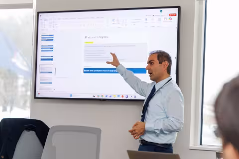 A presenter gestures to a projected slideshow with the title "Practice Examples" during the 2026 AI Plus Symposium.