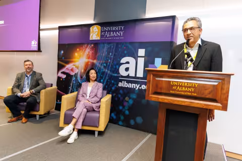 AI Plus Institute Director Balakrishnan (Prabha) Prabhakaran speaks at a podium while Provost Carol Kim and another speaker seated beside him on stage listen during the 2026 AI Plus Symposium.