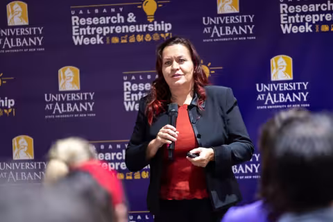 A woman in a black suit standing in front of a purple backdrop with the words "Research and Entrepreneurship Week" while holding a microphone a slide clicker. 