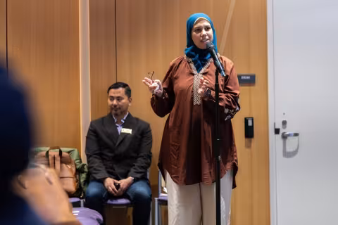 A woman stands at microphone speaking to someone out of frame while gesturing with her right hand. 
