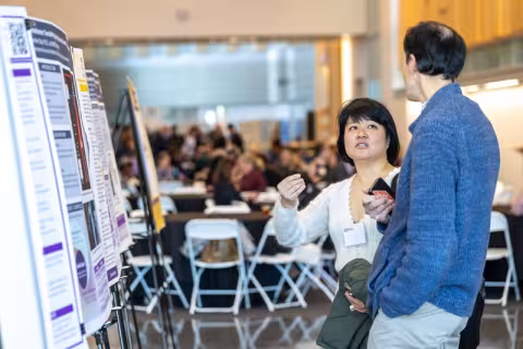 A man and a woman stand in the foreground discussing a poster in front of them with a large crowd seated at tables behind them.