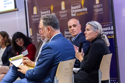 Six people sitting in a semi-circle on stage in front of a purple banner with the words "Research and Entrepreneurship Week" on it as one woman in a black sweater holds a microphone and talking.