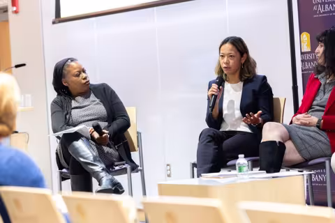 Three women seated on a stage; the one in the center is holding a microphone speaking while the others to her left and right look at her.