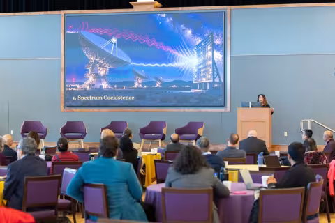 A person stands at a lectern on stage in front of a crowd gesturing to a blue-hued screen with a large satellite dish on it.